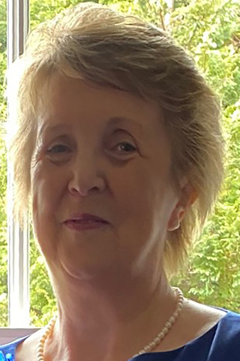 Headshot of a smiling Jill Clark, counsellor, an older woman with short blonde hair, wearing a royal blue dress and a white pearl necklace and earrings stands in front of a window with a hedge outside.