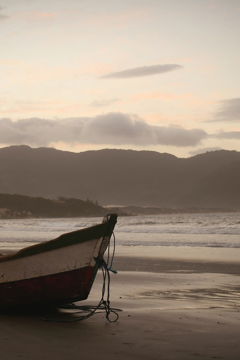A brown and white wooden rowing boat on a sandy beach, with waves gently lapping the shore. The sun sets over dark hills in the background.