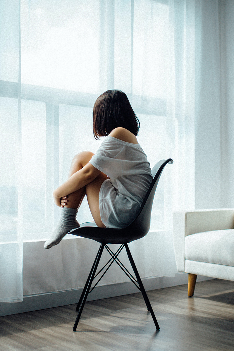 Tranquil-Counselling-By-Jill-Whitehills-Banff-Aberdeenshire-Isolation-Depression Dark haired woman wearing a white t-shirt and grey socks, sitting on a black chair in front of a window with a net curtain.
